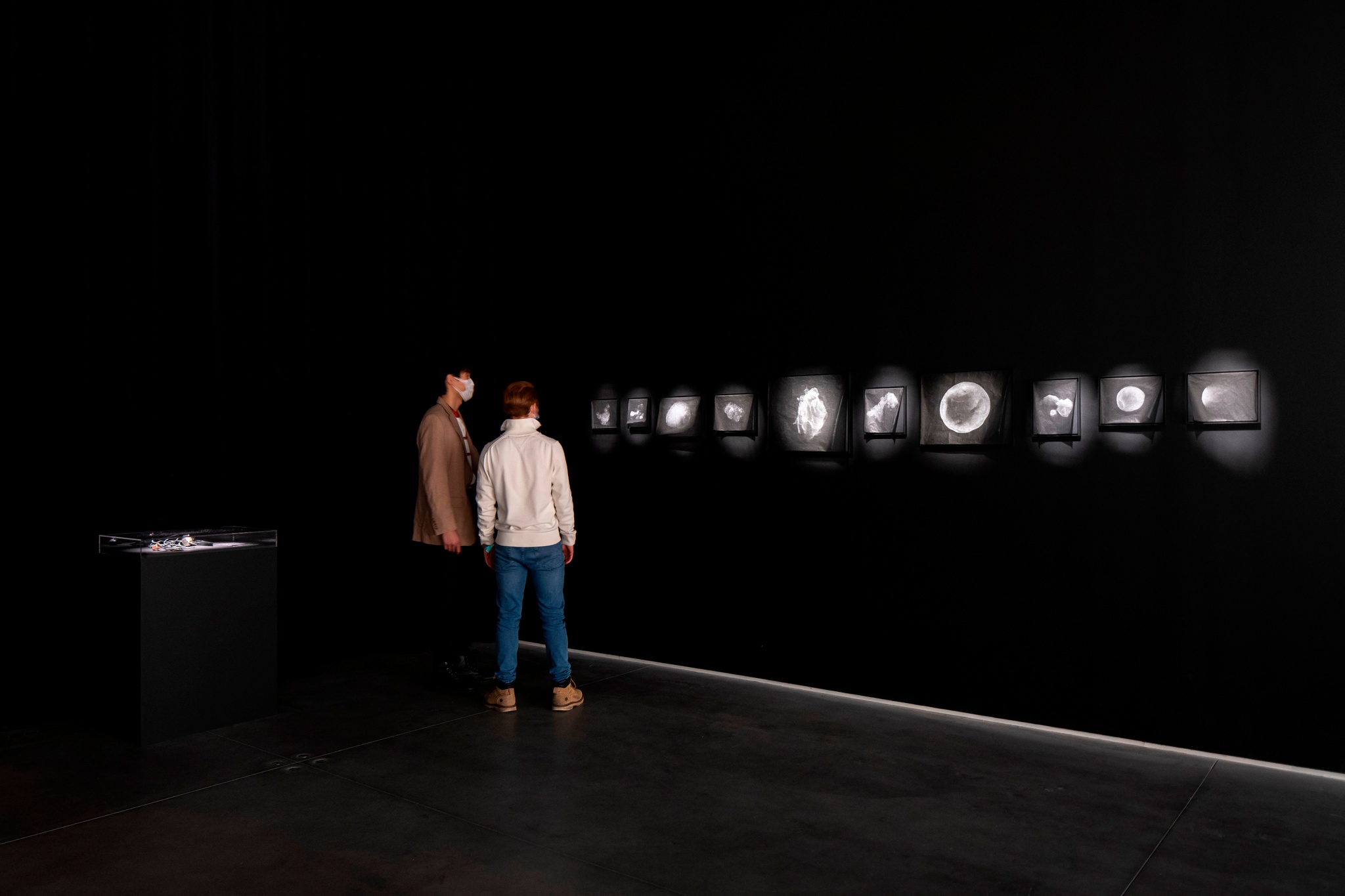 Two people stand in a gallery to the left of a series of ten photo prints on the wall. They are printed on thin black paper and depict particle pollution blown up as if under a microscope and recall images of stars.