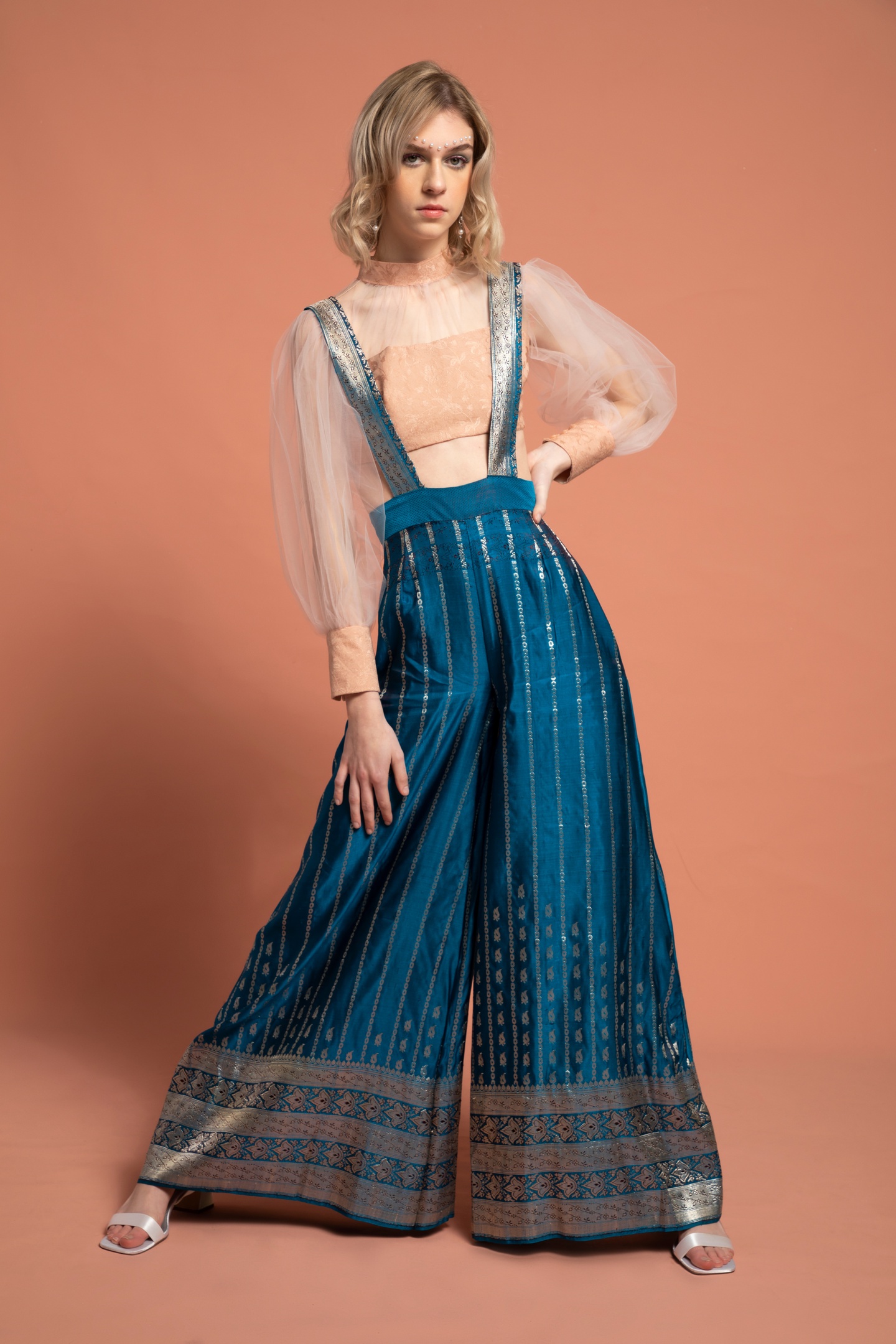 Model wears high-waisted overalls with extremely wide legs made of blue silk with a golden vertical stripe imprint and a wide border design at the cuffs. The top is a long sleeve crop top with peach fabric across the bust and at the collar and cuffs, connected by gauzy fabric over the chest and sleeves.