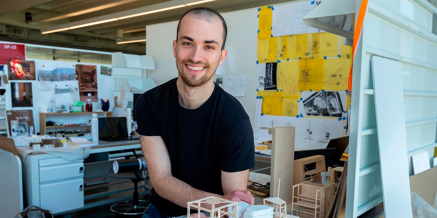 Portrait of Nathan Stanfield in his architecture studio. He sits at a white metal desk surrounded by sketches and wooden models. 