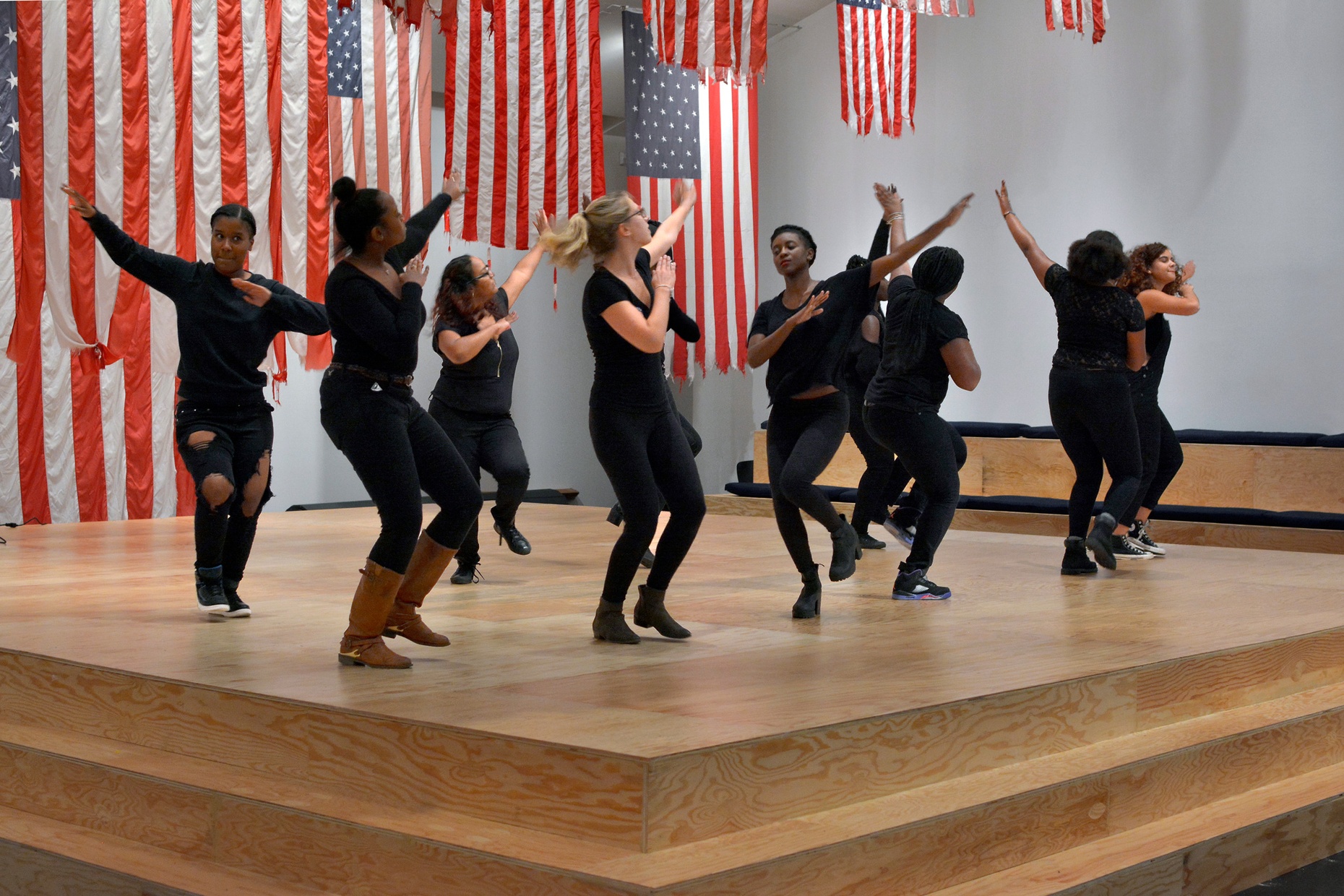 A group of people wearing black dance on a wooden stage surrounded by American flags hung from the ceiling.