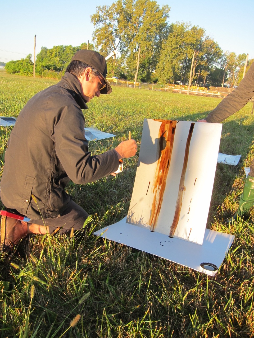 Shaun on his knees painting brown lines on a rectangular canvas which is held upright by someone else