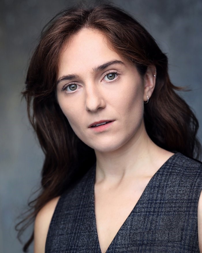 A headshot of actor Jessica Revell, who looks directly at us with head cocked to the side. Jessica is white with long dark auburn hair. She wears a gray vest. 