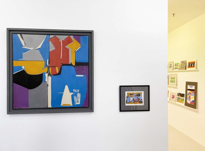 Gallery installation featuring one large abstract work in blues, gray, gold, purple, black, and red on the left side of a gallery wall, with a smaller piece to its right; in the background, one can see further into the gallery, where more works are hung on the walls.