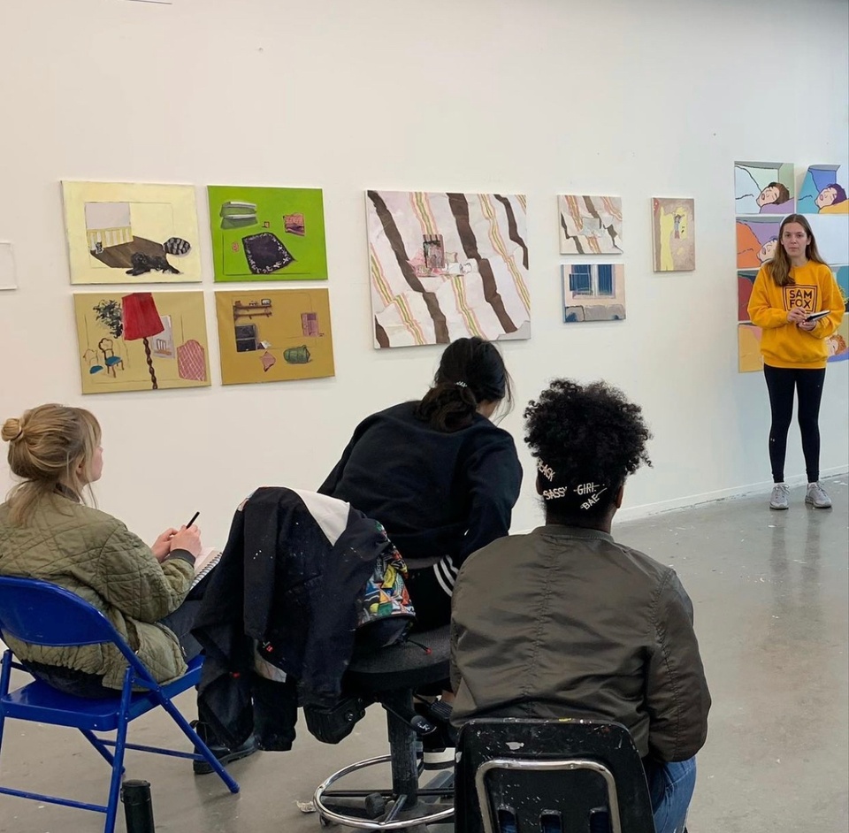 A student wearing a Sam Fox jumper presents her paintings to the class. The colourful paintings depict household furniture floating in the scene in plain colorerd backgrounds. 