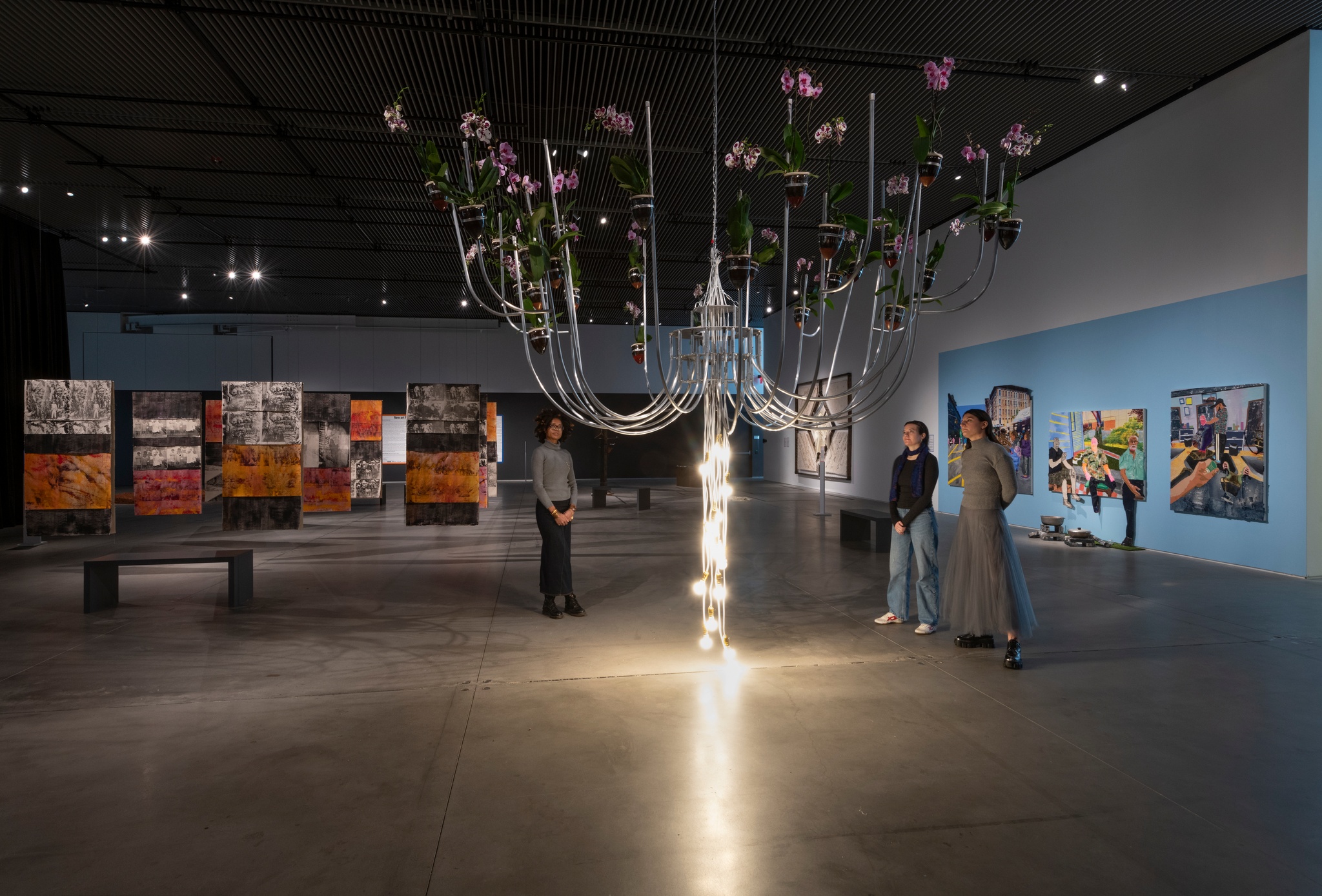 Three gallery goers stand around a sculpture that hangs from the ceiling. It resembles a chandelier with long aluminum pipes bent upward gracefully. At the end of each pipe hangs an orchid in a ceramic pot. From the center of the sculpture, a string of LED light bulbs hangs to the ground.