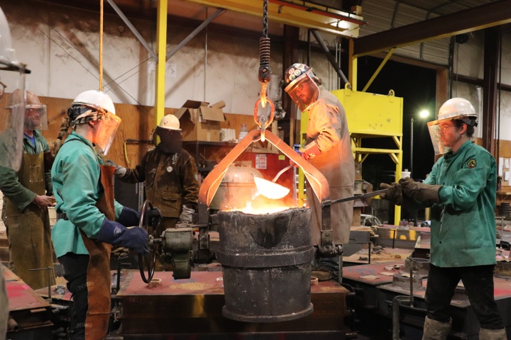 Two people in PPE watch as a third holds a massive ladle-like object near a vat of hot melted steel.