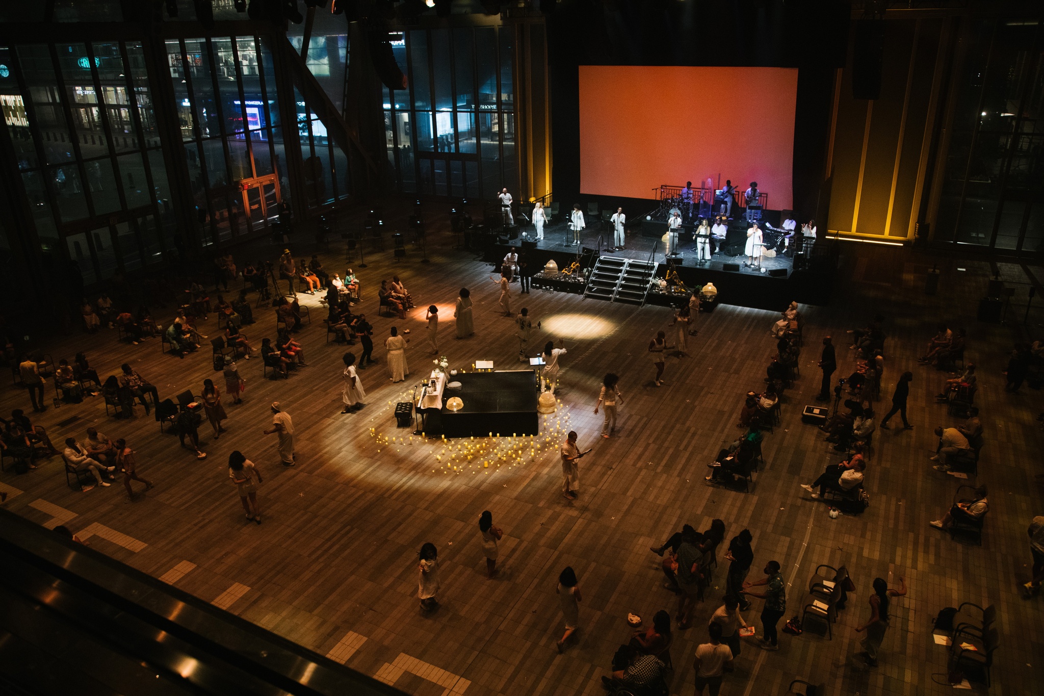 A piano and small black stage in the center of a large space with candles arranged in a circle around them. Performers line either side of the space with audience seated in shadow. A line of performers stands on a larger stage at the top of the space.