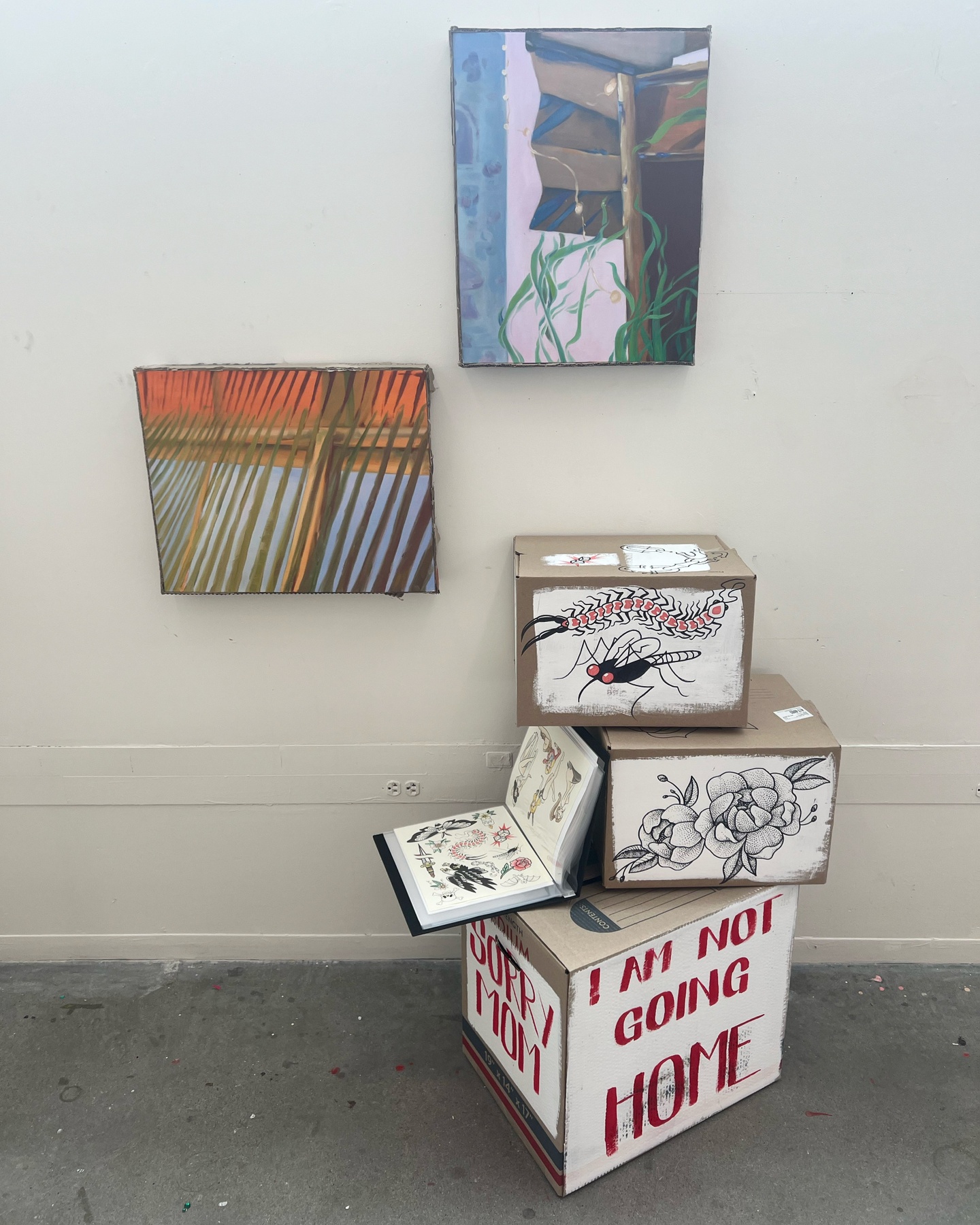 two paintings of exteriors displayed with a stack of painted boxes on the floor.