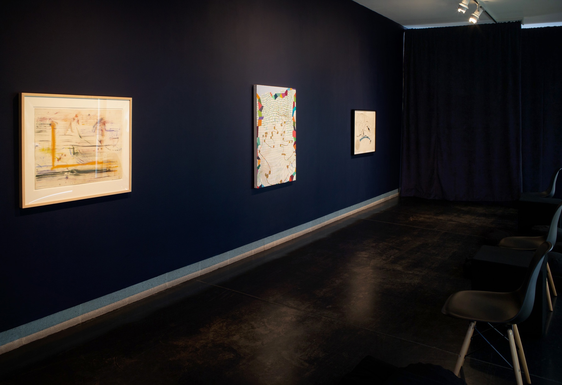 Three abstract artworks hang on a navy blue wall in a dark room with multiple black chairs facing the art.