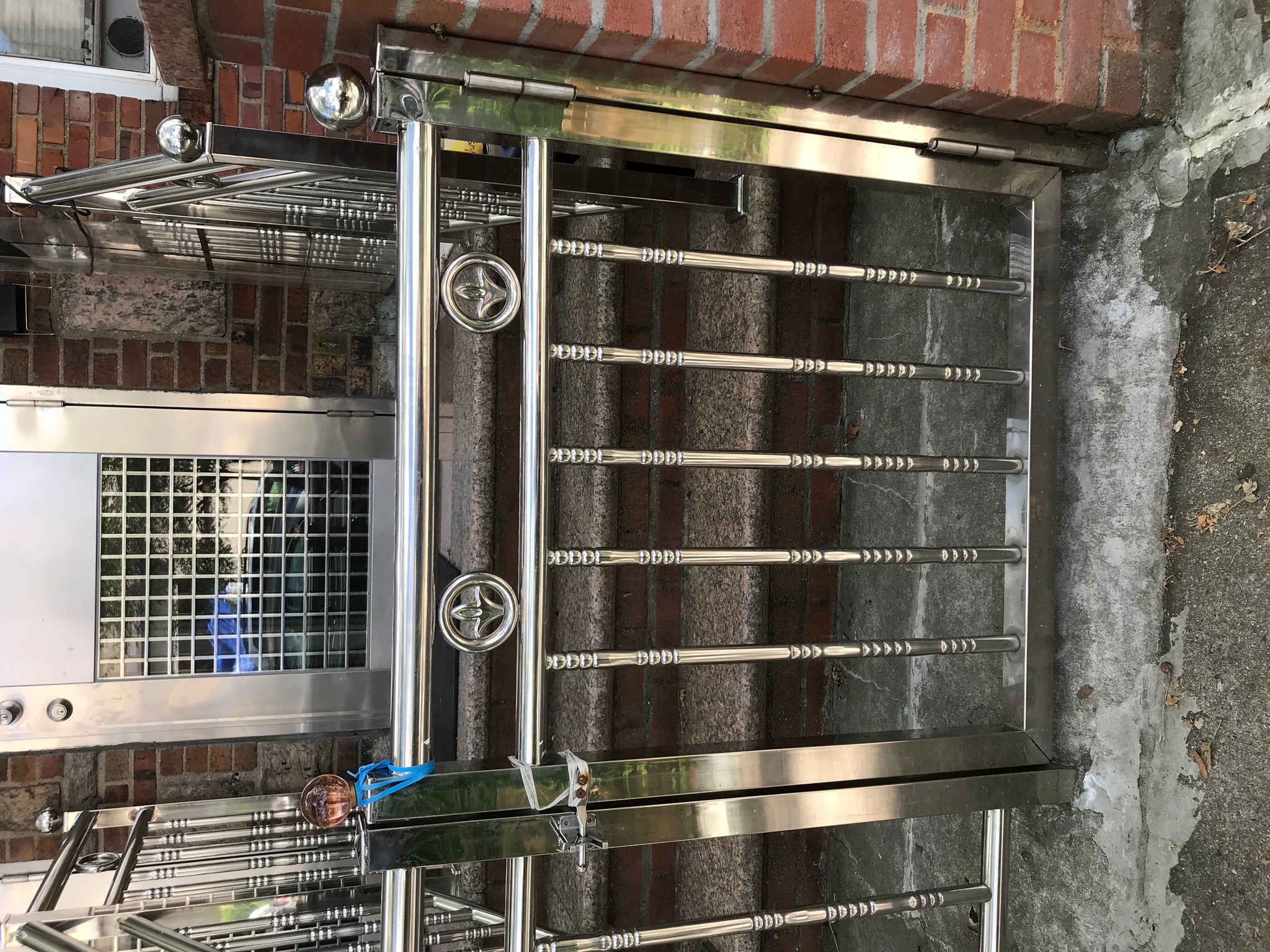 A shiny stainless steel gate with behind it up three marble stairs, a shiny stainless steel banister and railing leads to a stainless steel front door of a brick house