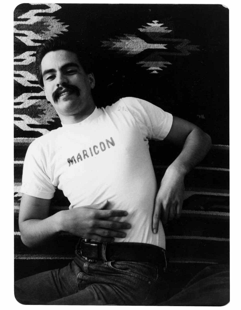 A black and white photo of a Latino man smiling laying on a blanket wearing a shirt that says "MARICON."