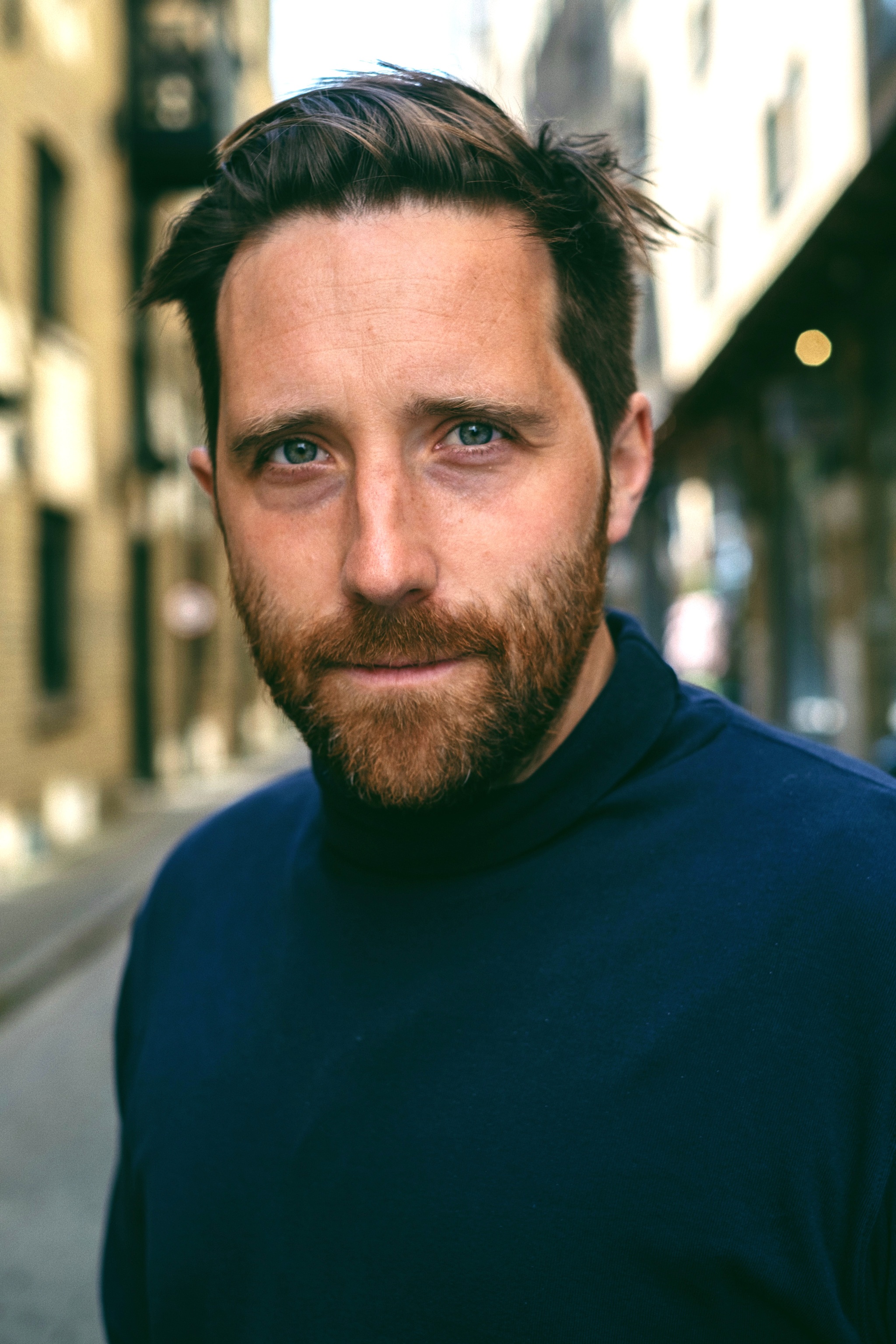 A headshot of actor Joseph Kloska, who looks at us intently. Joseph is white with dark brown hair, and a reddish brown beard. He wears a dark blue sweater. Behind him is a narrow street in dappled sunlight. 