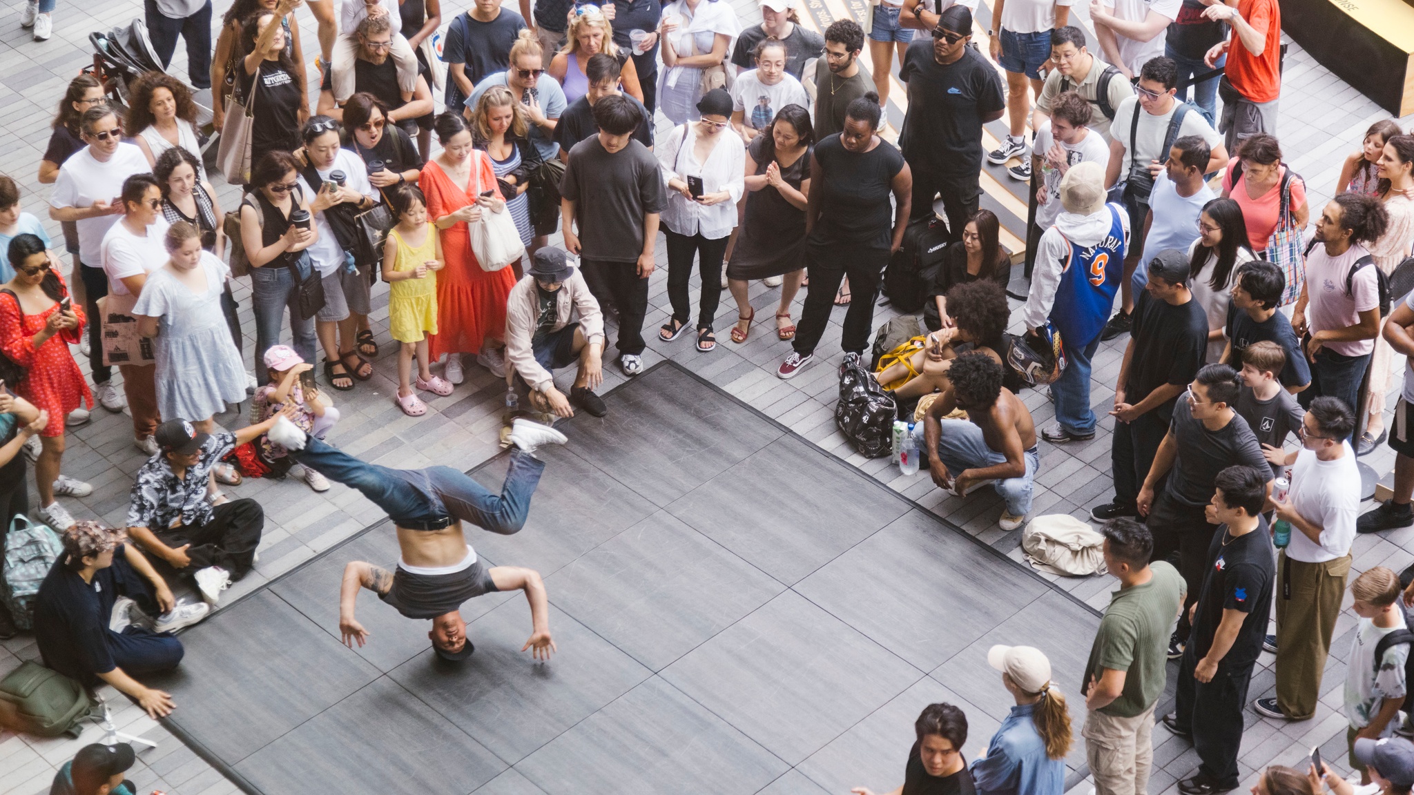 A breakdancer spins on his head on an outdoor dance floor surrounded by an excited audience. 