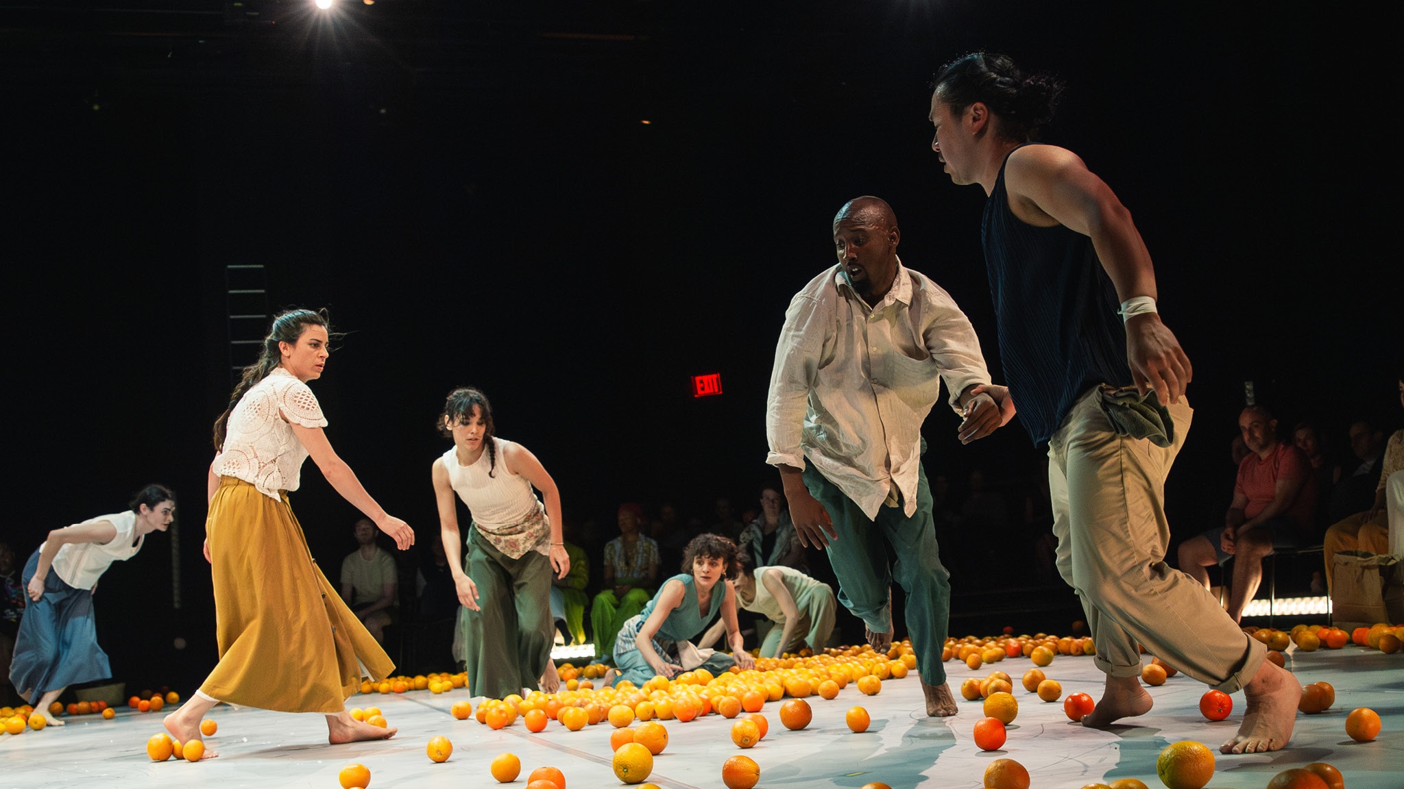 Dancers move energetically around a central performance space strewn with brightly colored oranges. 