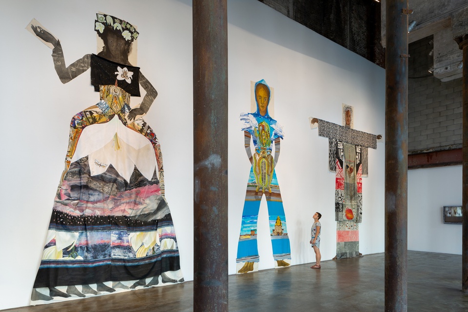 Photo of three large-scale 2D installations of figures on the wall of a gallery, with an individual gazing up at them.
