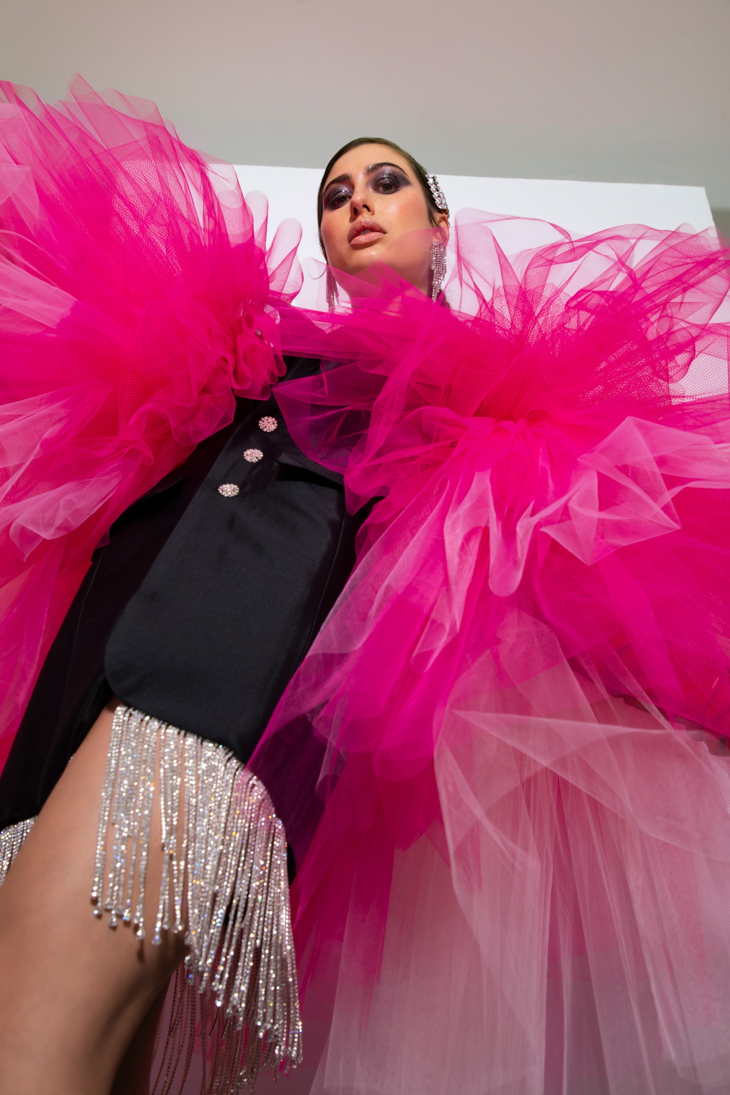 Model photographed from a low angle wearing a black satin minidress with a rhinestone tassel fringe. The dress's sleeves are voluminous ruffs of magenta tulle, each larger than the minidress.
