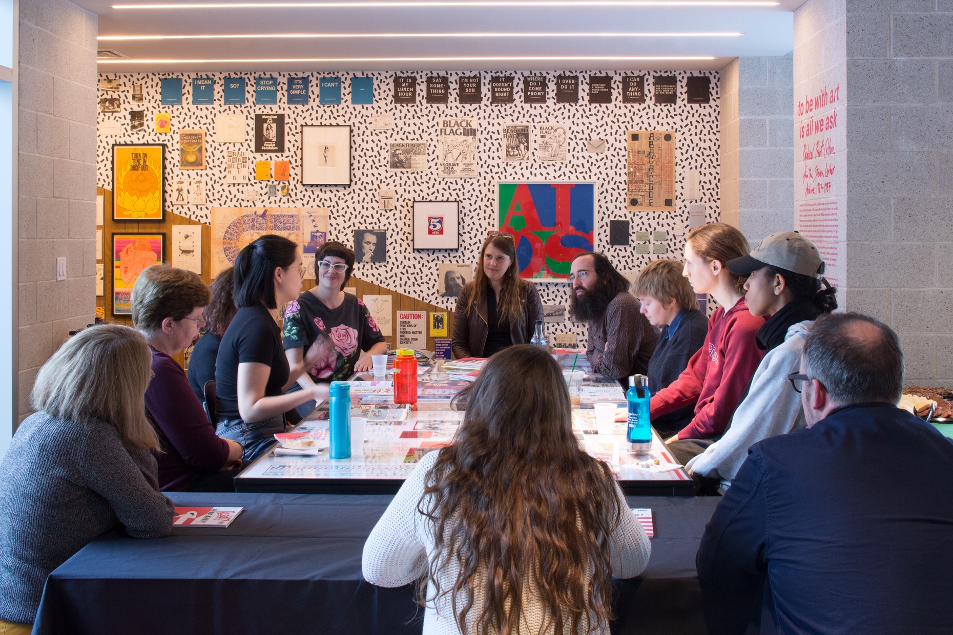 A group of people sit around a table covered with screenprints in front of a black and white polka-dot wall with colorful prints hung on it.