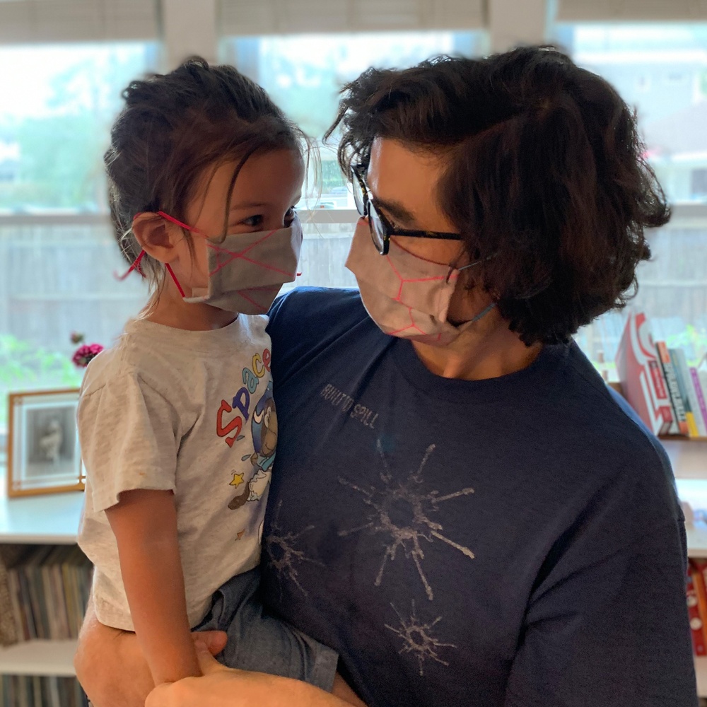 A light-skinned, dark-haired, young man wearing glasses wearing a pink and gray mask looking at and holding a young, light-skinned boy wearing a similar mask.