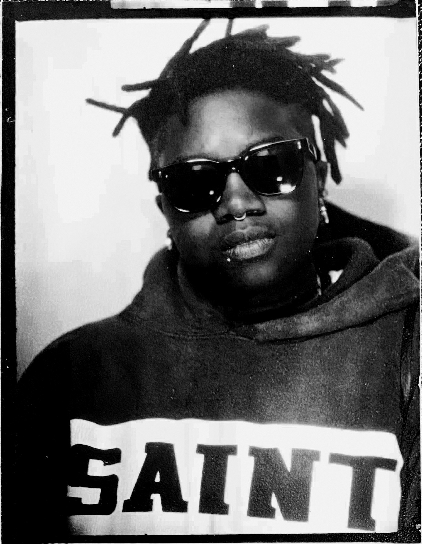 A black-and-white portrait of artist Tiona Nekkia McClodden wearing black sunglasses and a hoodie that reads SAINT in black letters across a white rectangular field