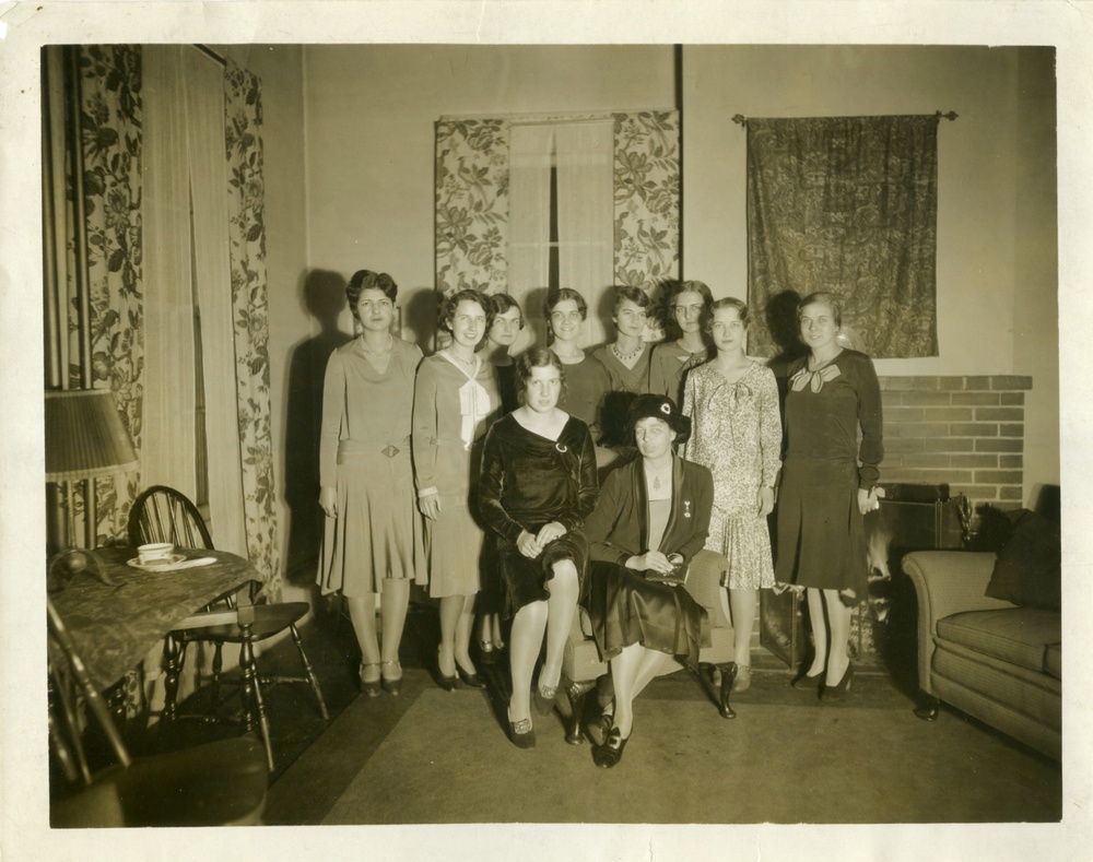 A sepia photograph depicts a group of standing light-skinned young women who are smiling at the camera and positioned around the former First Lady Eleanor Roosevelt seated in an armchair.