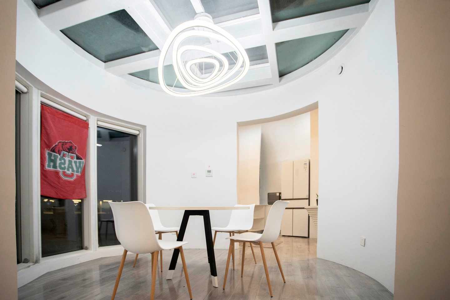 Interior of the house. White walls are curved; the left side of the room features three tall windos. In the center is a table with four white chairs with wooden legs. Overhead is a bright white light composed of irregulrarly shaped rings.