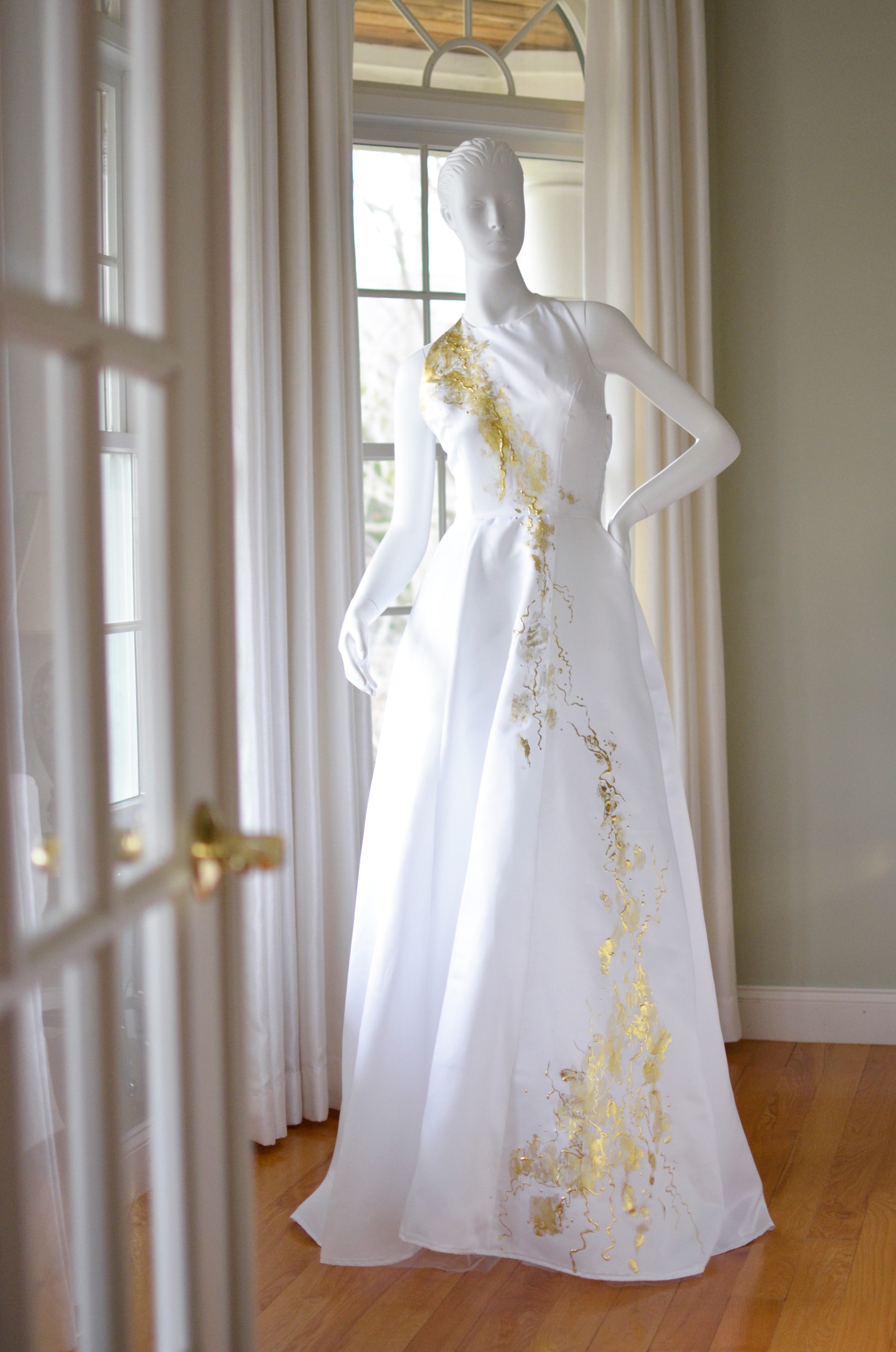 White floor-length sleeveless dress with gold paint applied in a streak from the right shoulder across the front of the dress down to the floor.