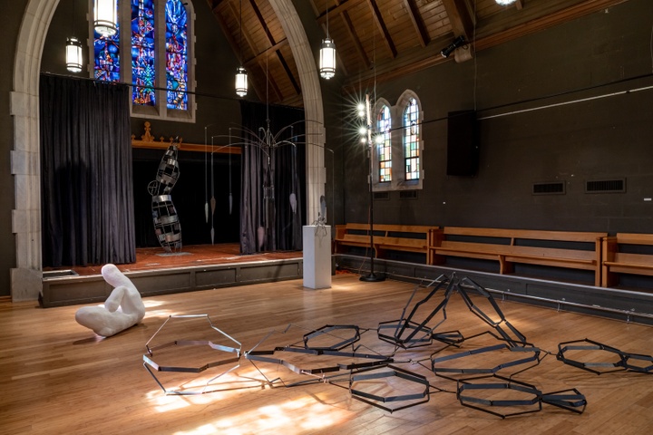 Chapel space with an arched ceiling and stained glass windows displays various artworks.