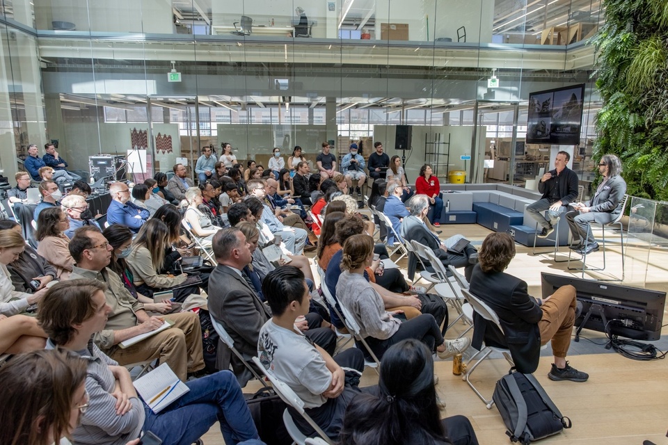 Indoor courtyard with a living green wall is filled with seated students and faculty attending a symposium. Two lecturers sit in front of the green wall in conversation. On a screen behind them is a procedurally-generated architectural image.