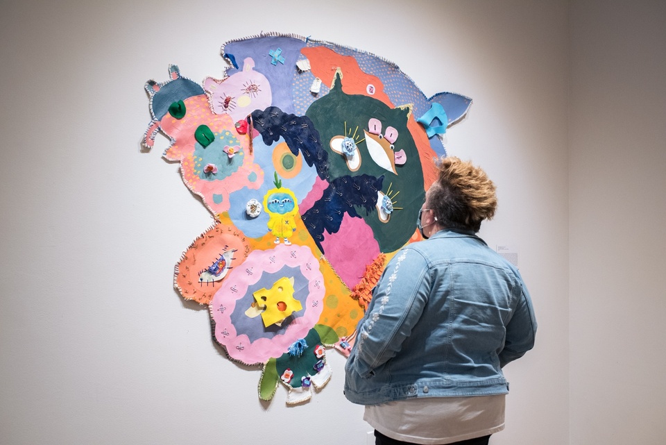Person looks at a brightly colored fabric wall hanging. The design is of many whimsical, cartoonish characters in bright pastels.