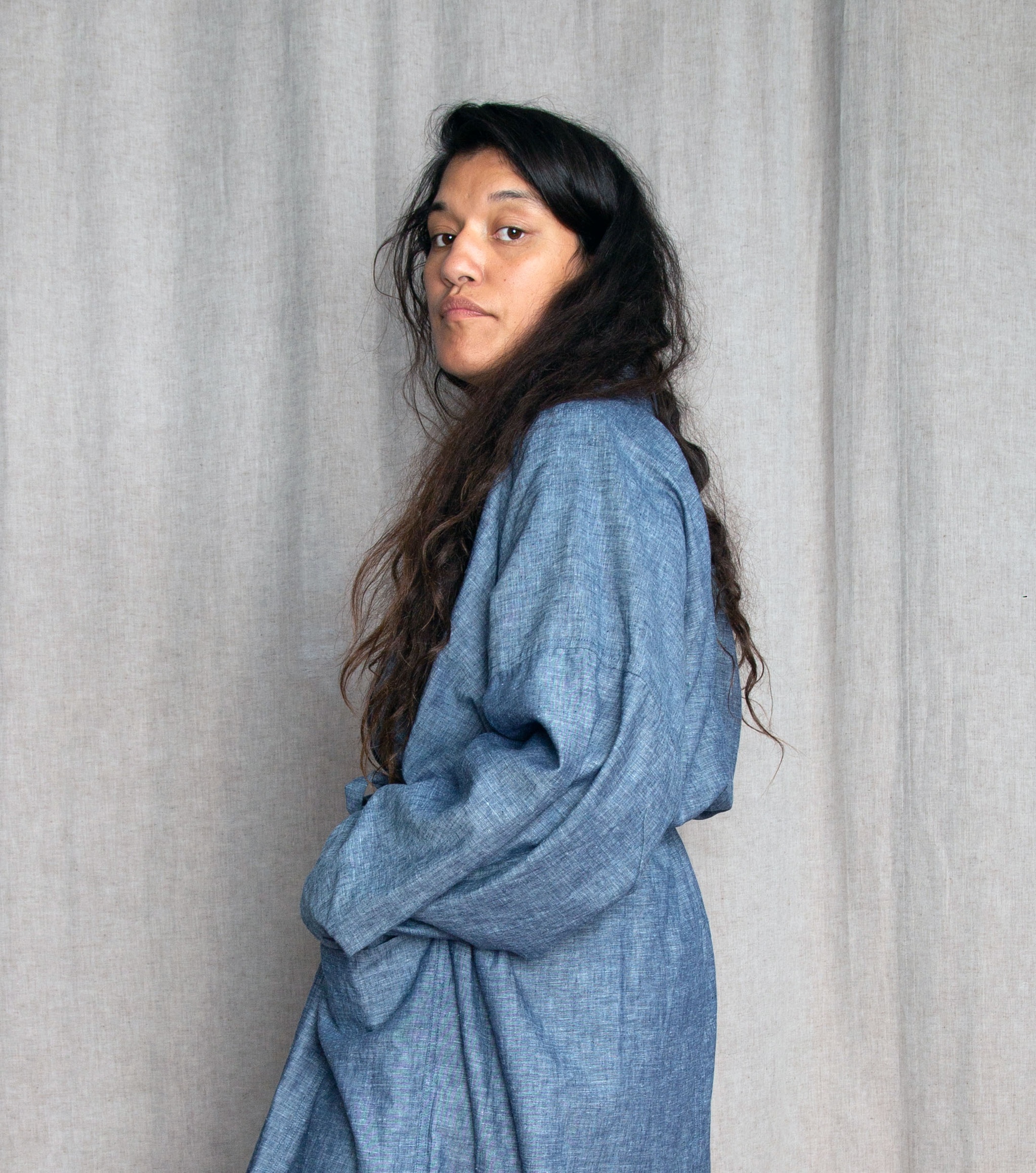 The artist Pelenakeke Brown stands with her shoulder and face turned to the camera. She has long brown hair that falls over her shoulder and back and holds one hand to her waist. She wears a draped, denim-colored garment that resembles a dress or robe. 