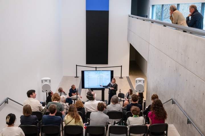 View from the back of the event space in the Pulitzer showing a crowd seated on risers, facing the panelists, and a few attendees watching from the balcony to the right.