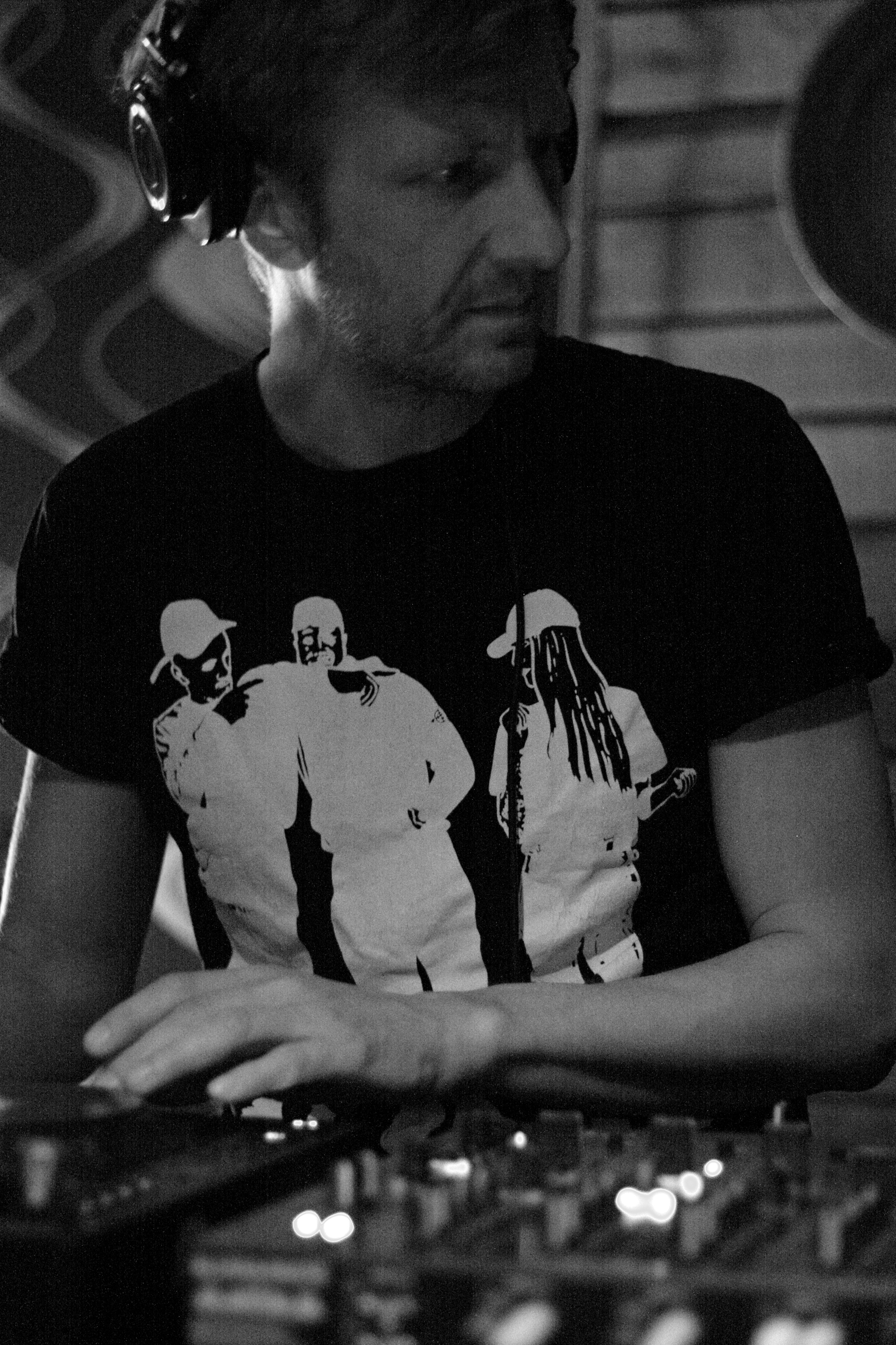 A white man in a black tshirt with a graphic depicting three people dressed all in white looks to his left while working a DJ booth. He wears headphones over his ears. 