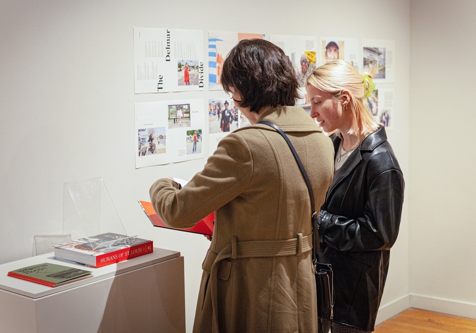 Two students look at a copy of the "Humans of St. Louis" photobook on a plinth. On the wall behind them are sample layouts from the book, tacked to the wall.