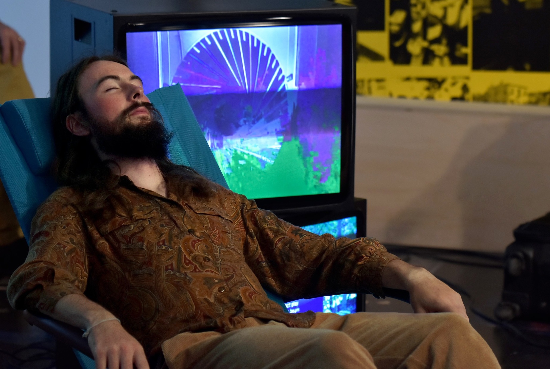 A light-skinned, young man in a tan, paisley shirt reclines in a cushioned blue chair with his eyes closed in front of two TVs stacked on top of each other displaying abstract patterns.