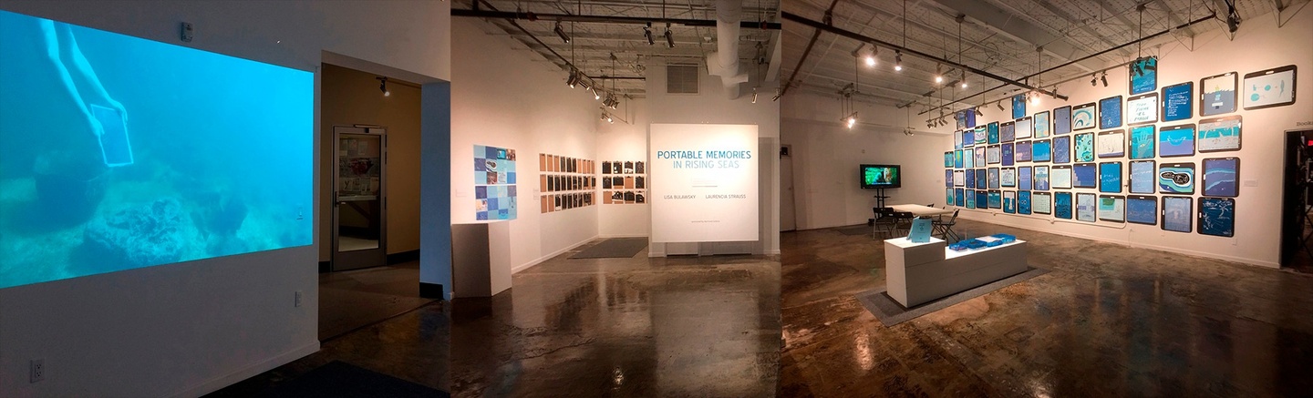 An exhibition in a white gallery with dark floors. In the center of the image against a wall/panel is the title "PORTABLE MEMORIES IN RISING SEAS" in blue. On the walls are artwork, as well as video projections in the front and back. Sculpted objects are also on a white platform in the center of the space.