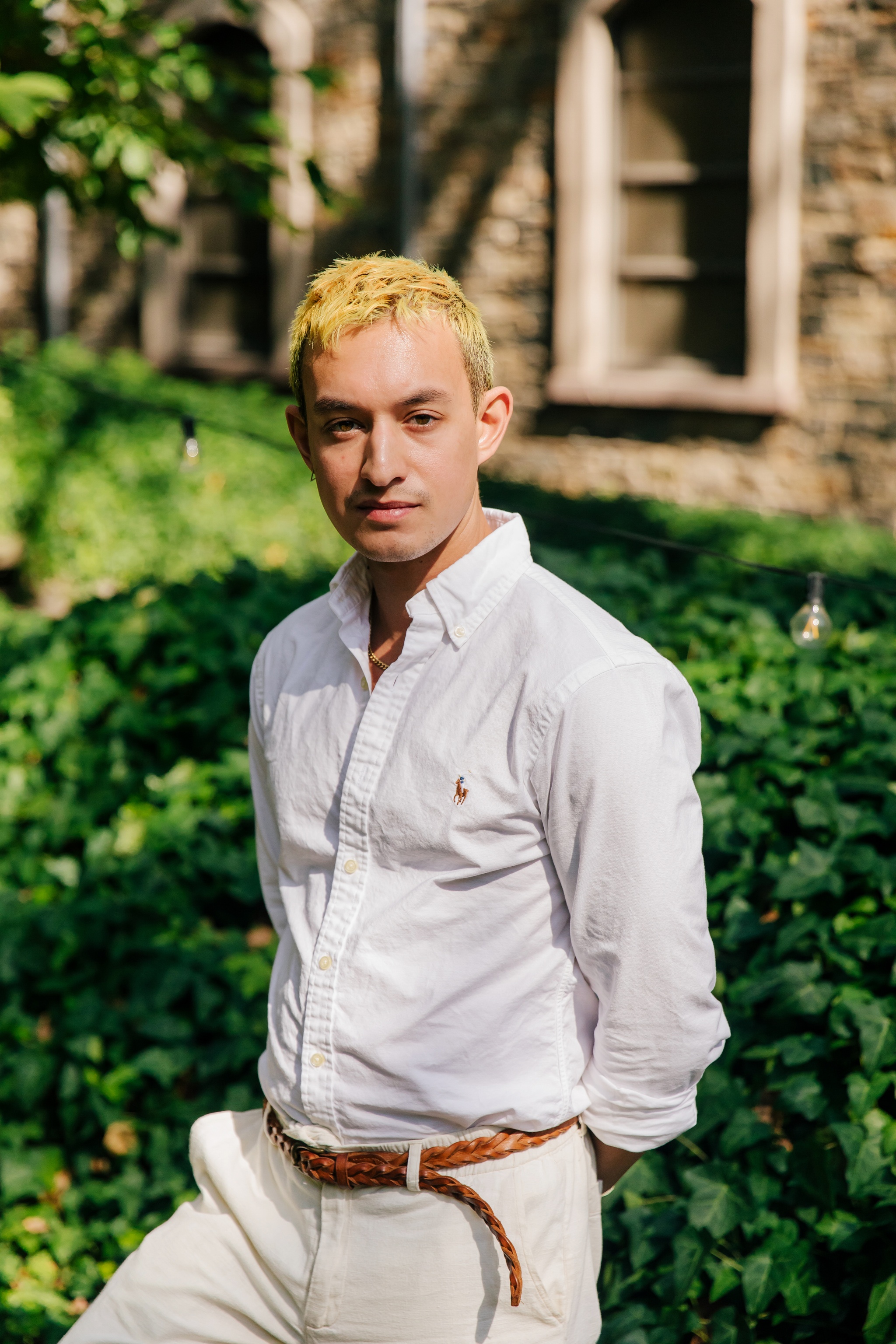 A portrait of Kyle Dacuyan. He stands outside in a garden area with green bushes behind him and a stone building in the background. He poses with his hands behind his back. He wears a white button down shirt, white slacks, and has bleached blond hair. Photo by Tess Mayer