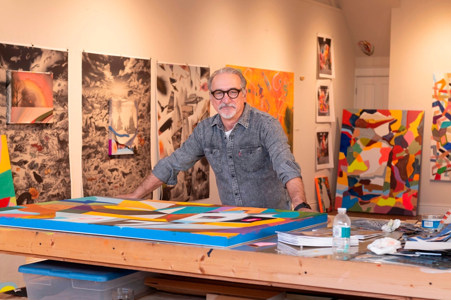 Carmon Colangelo resting hands on a desk in his studio with art on the walls behind him