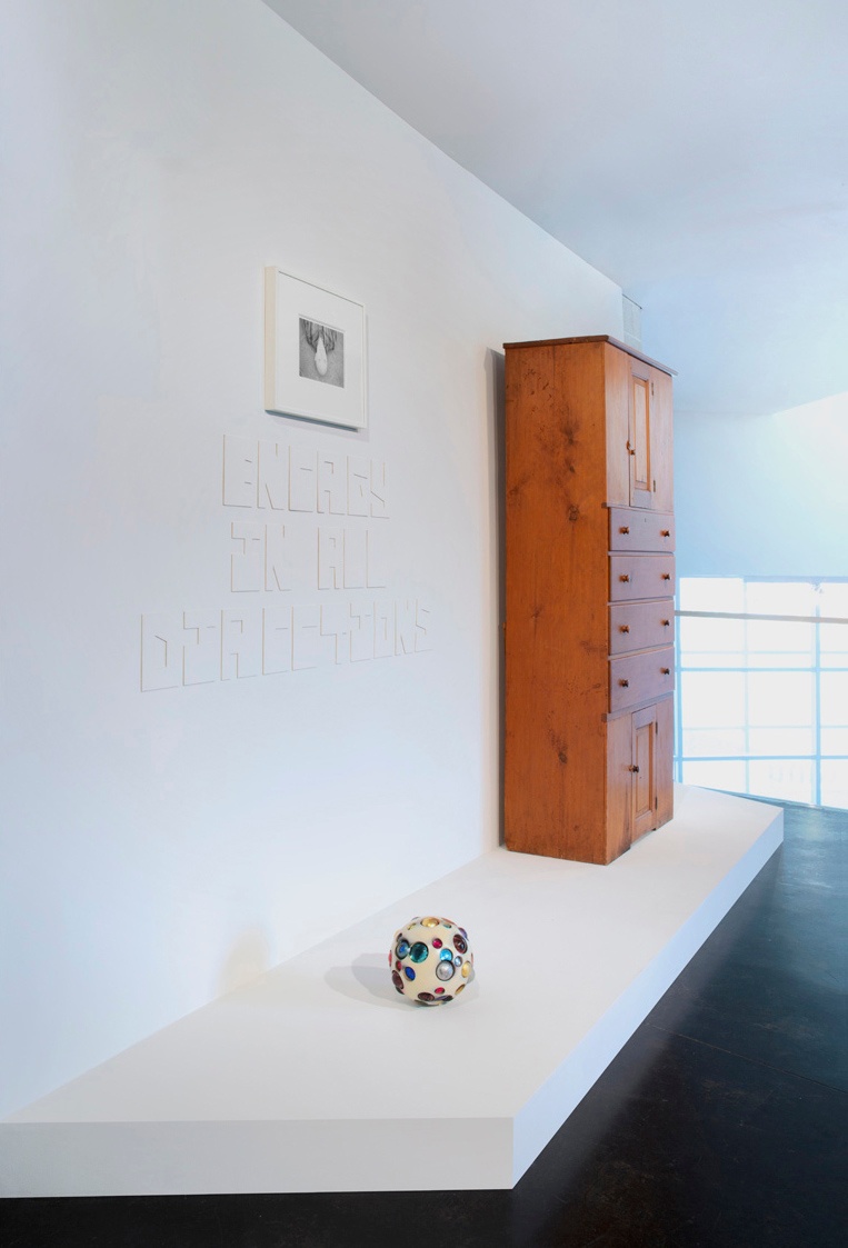 A tall, wooden cupboard sits on a white plinth to the right of a small, white sphere covered in colorful circles. A black and white photograph and white text "Energy in All Directions" hang on the white wall behind them.