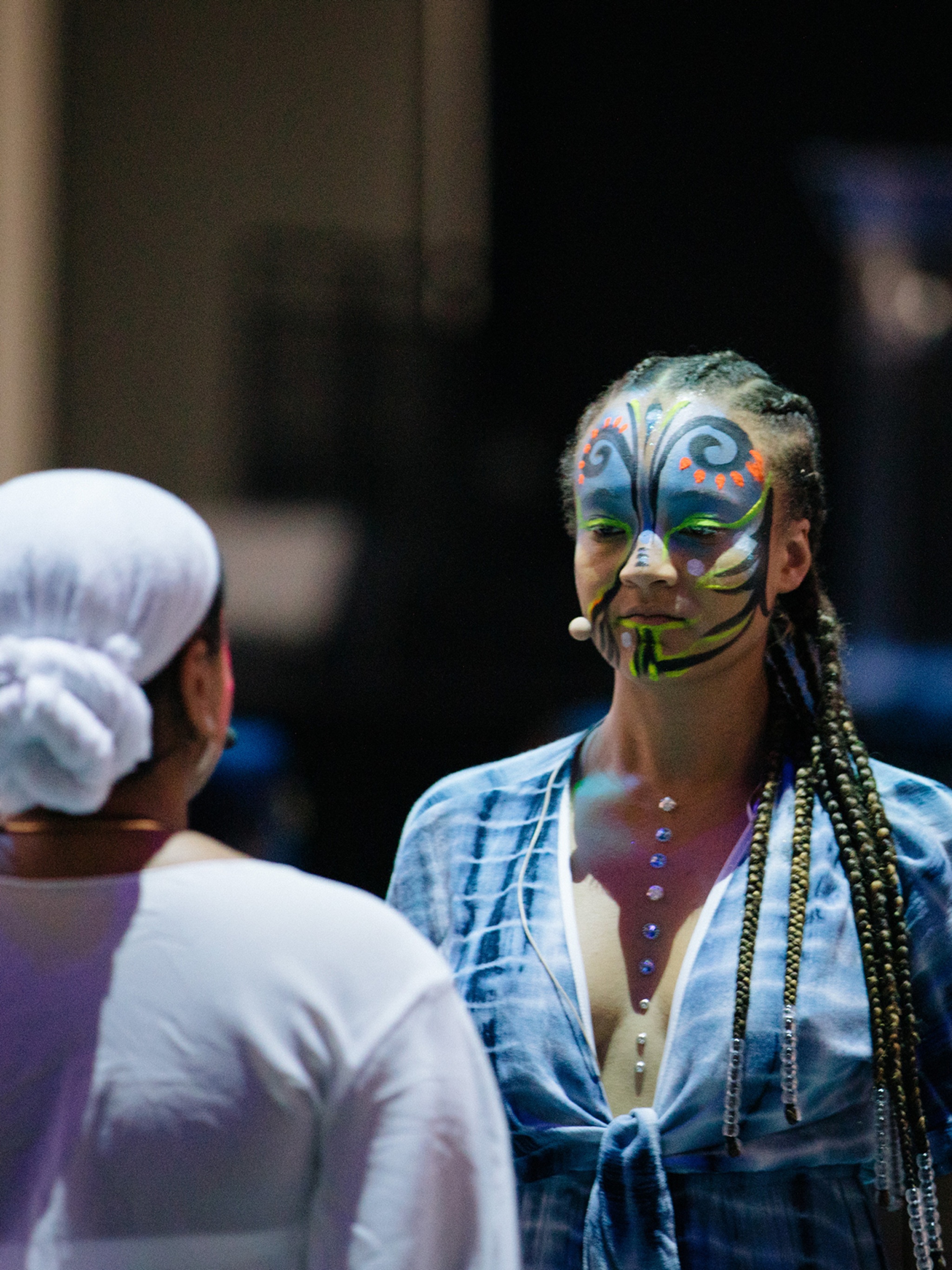 A person with face paint resembling a butterfly faces another performer who is seen from the back. 