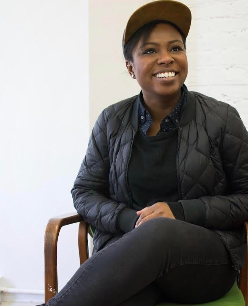 Lighting designer Stacey Derosier smiling, sitting with her hands together in her lap and wearing an all-black outfit and cap