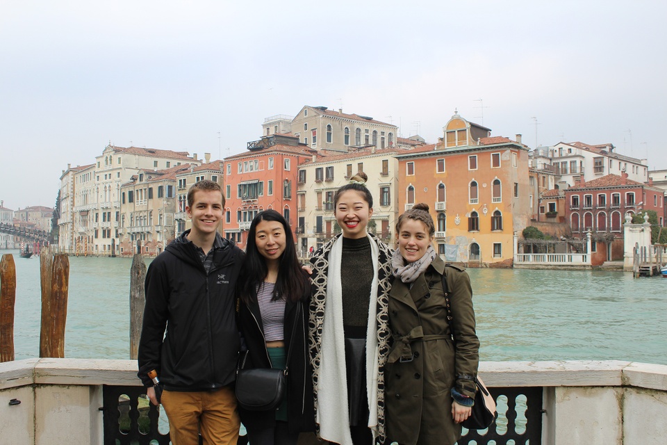 Four people in Venice standing in front of a blue canal with pink and yellow buildings behind them on a cloudy day.