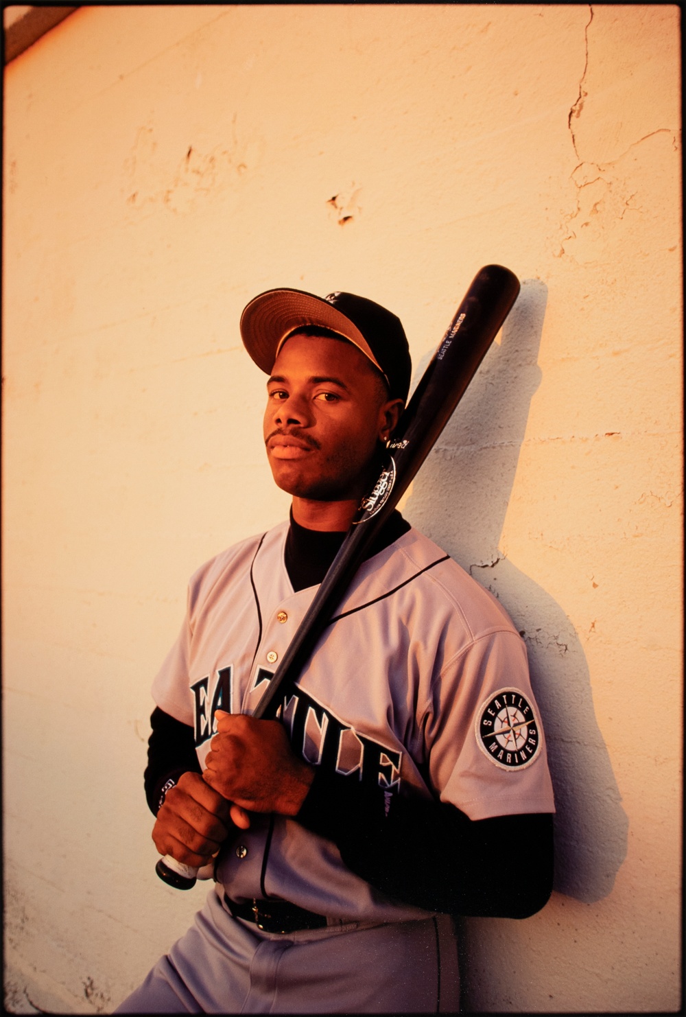 A man in a baseball uniform holds a bat and loos at the camera while leaning against a white concrete wall.
