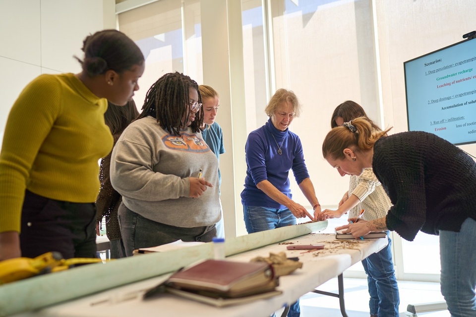 Students gather around a narrow green trough on a tabletop holding soil core samples while an instructor demonstrates the analysis.