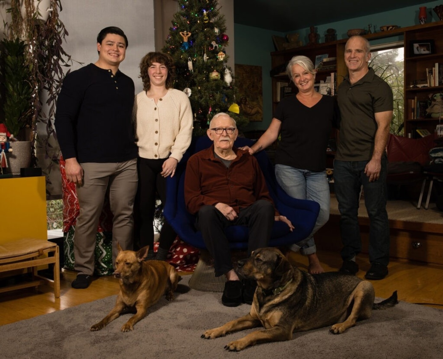 A man sits in a chair with two adults on either side of him. Two dogs are laying on the ground in the foreground.