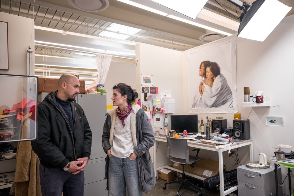 Two people stand in a studio space and chat. The studio contains a desk with a monitor and sound system, file cabinets, and a large square canvas with an image of two women embracing, wrapped in a white sheet.