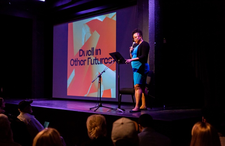 Person on stage presents to a crowd in front of a screen reading "Dwell in Other Futures."