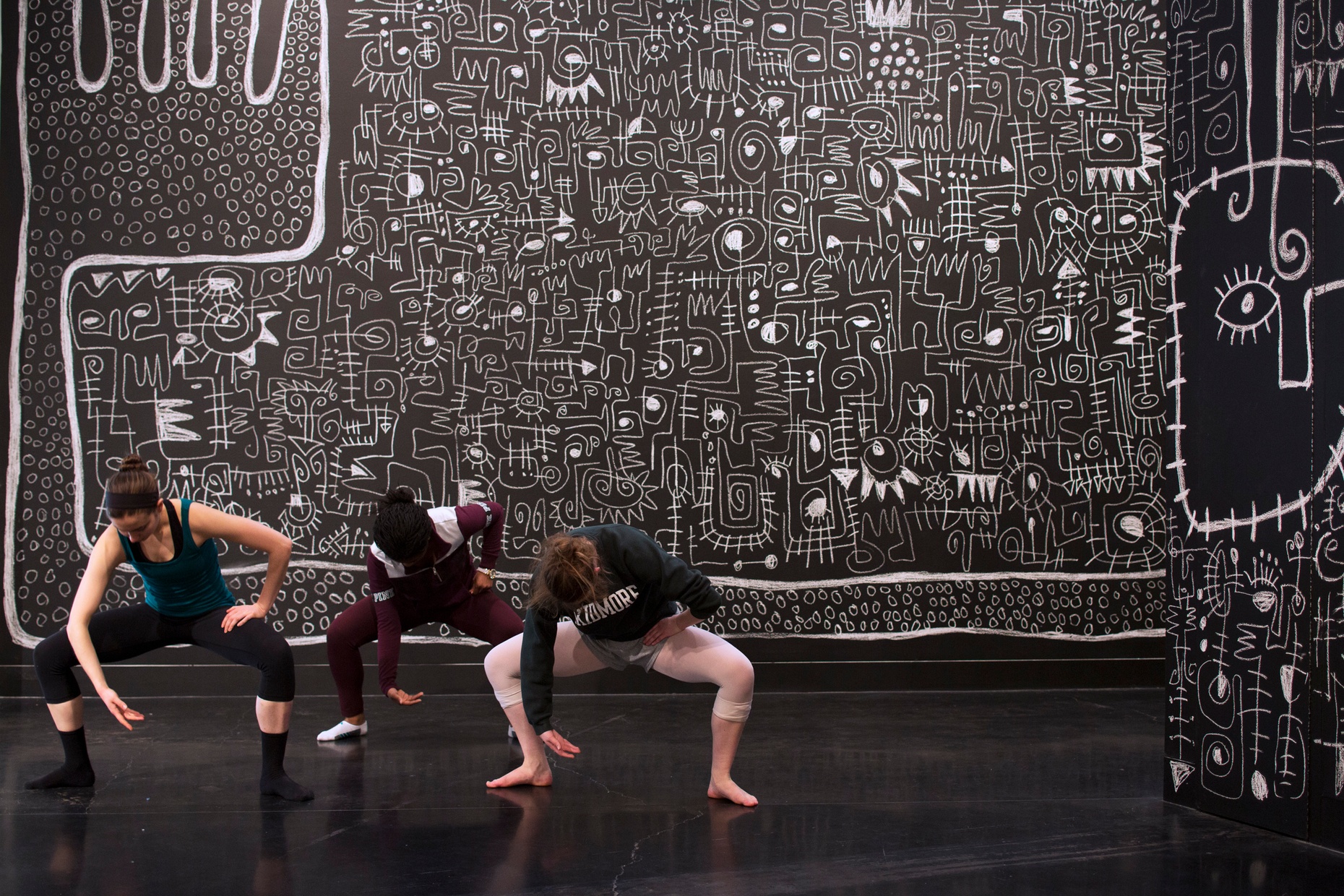 Three female dancers bend in a squat in front of a charcoal background covered in white doodles.