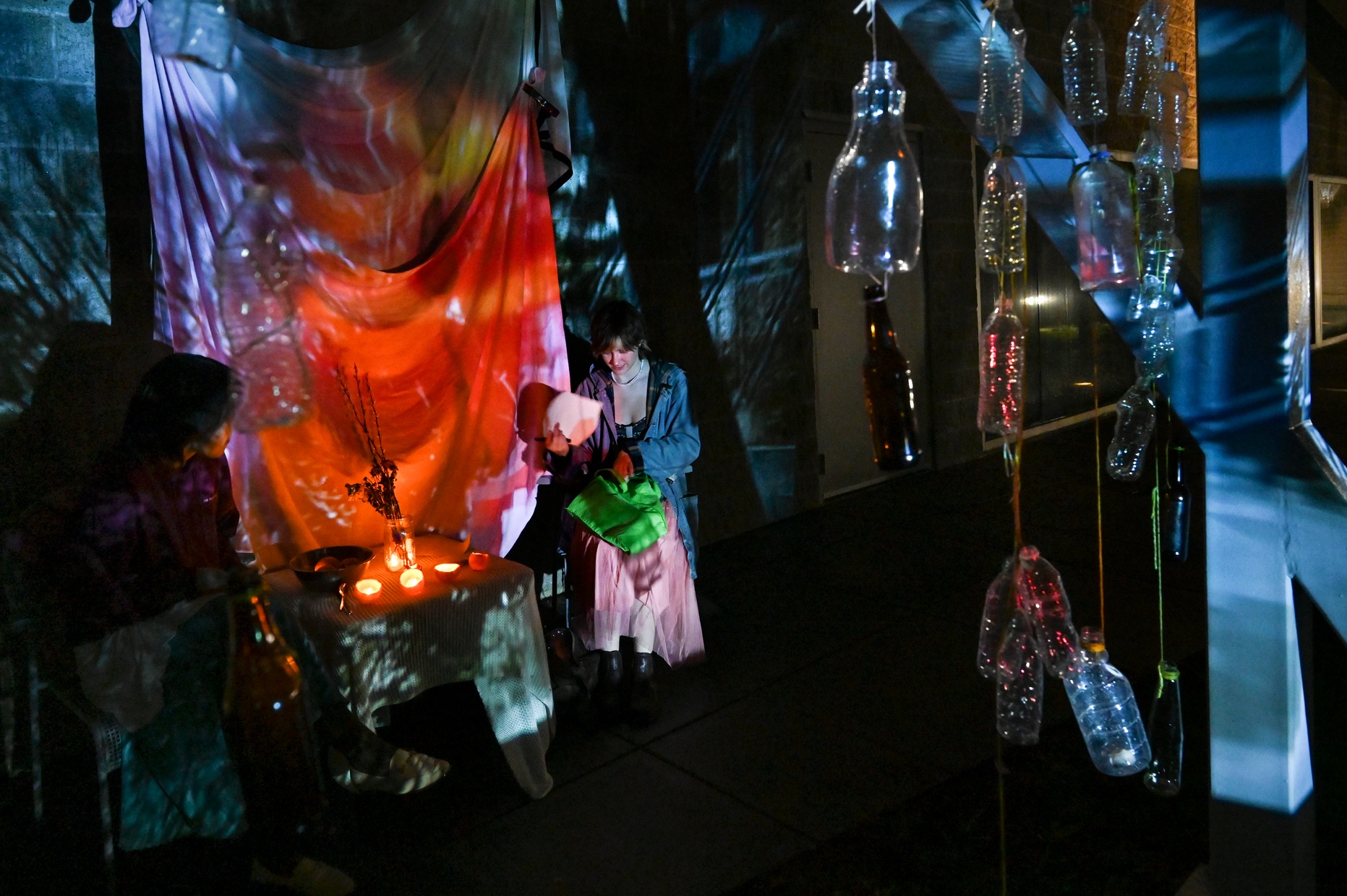 Two people sit in an art installation dimly lit with colorful lights and hanging bottles.