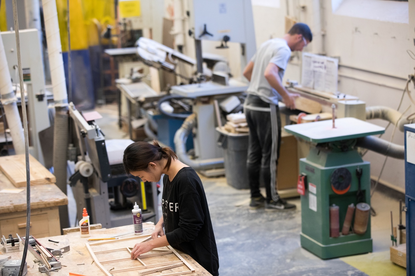 Two people working at opposite ends of a shop space featuring woodworking and welding tools.