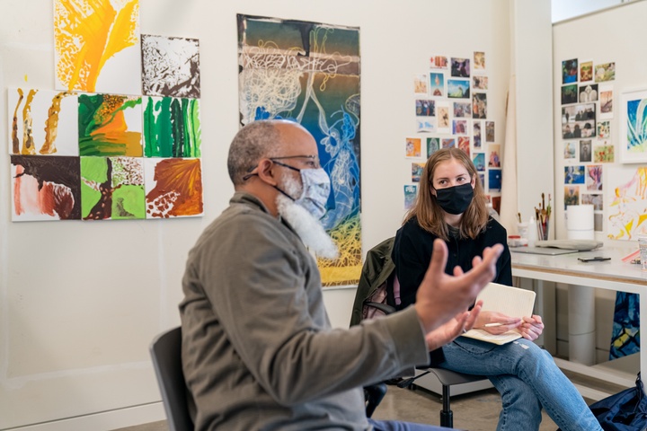 Two people chat in a studio space with drawings pinned on the wall behind a desk to their right.